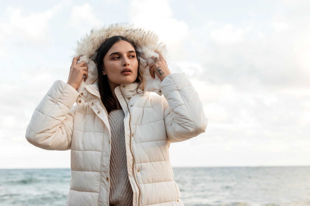 A woman wearing a white coat and hat stands by the ocean, gazing at the waves under a clear blue sky.