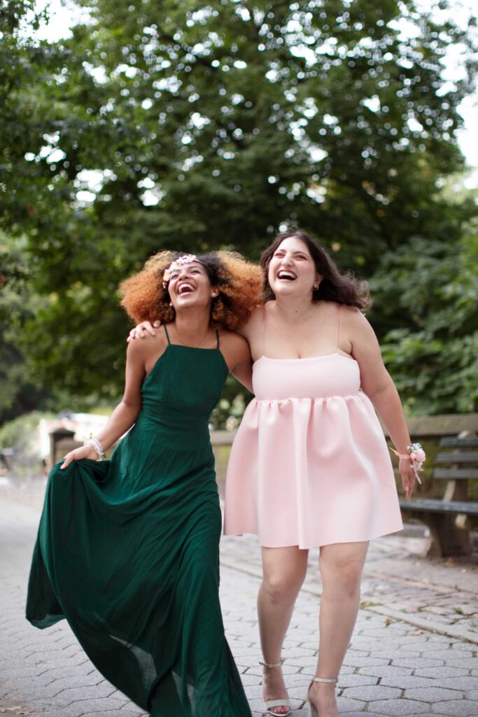 Two women in elegant dresses share a joyful moment, laughing and smiling together in a vibrant setting.