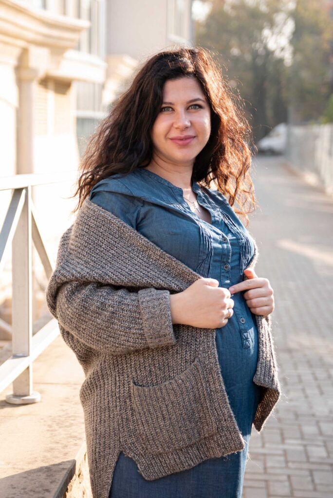 a plus size woman dressed in a denim dress and cardigan, lovingly holding her belly while exuding a calm demeanor.